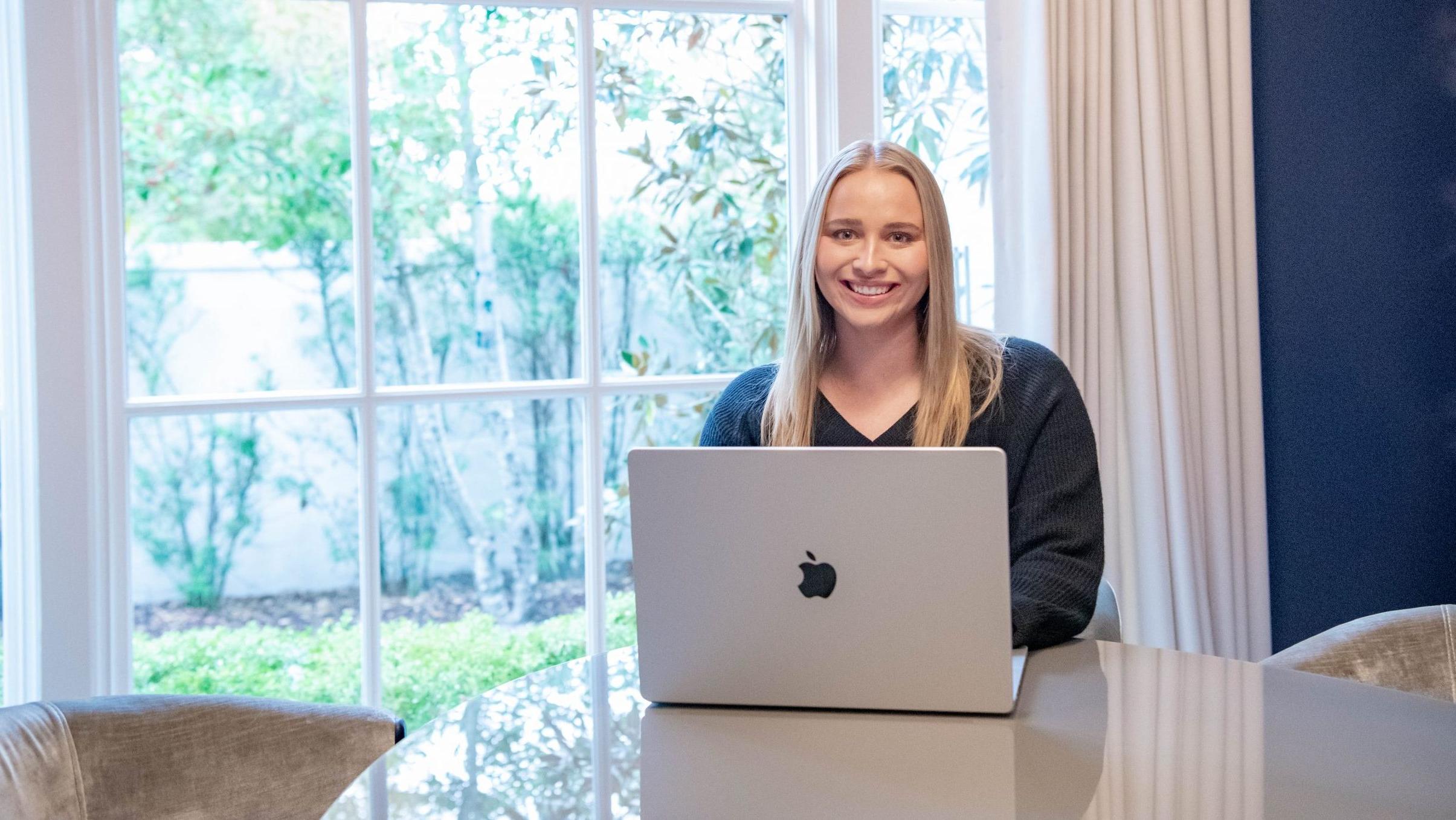 Student sitting at dining room table, with laptop and large window behind her at home.